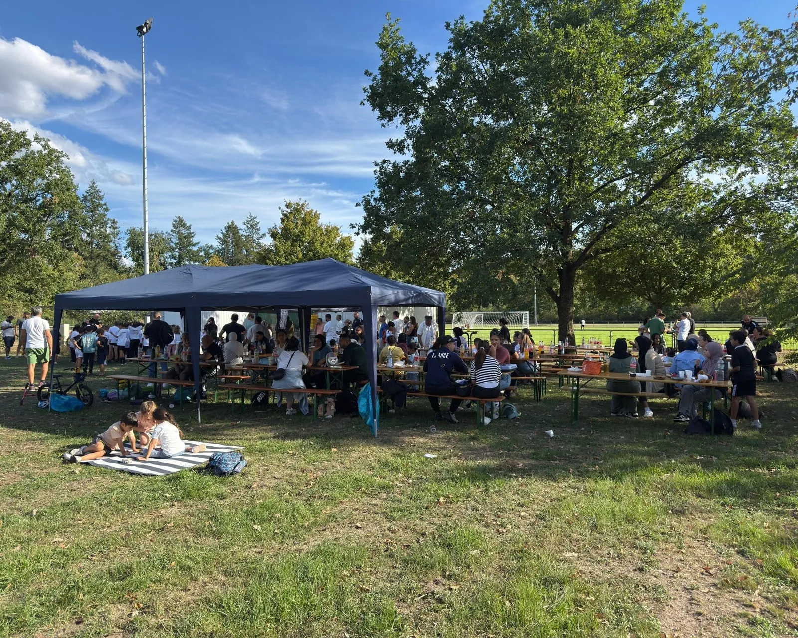 Pavillon mit Bierbänken und Gästen unter Bäumen neben dem Flutlichtmast