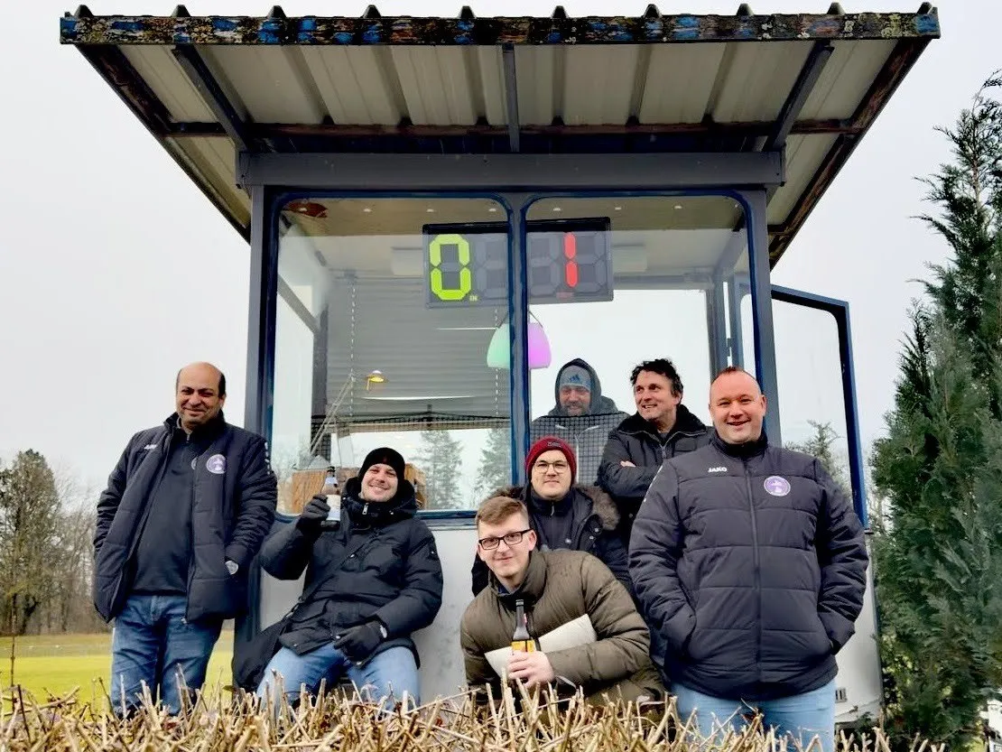 Vereinsmitglieder vor der Sprecherkabine mit Anzeigetafel am Spielfeldrand