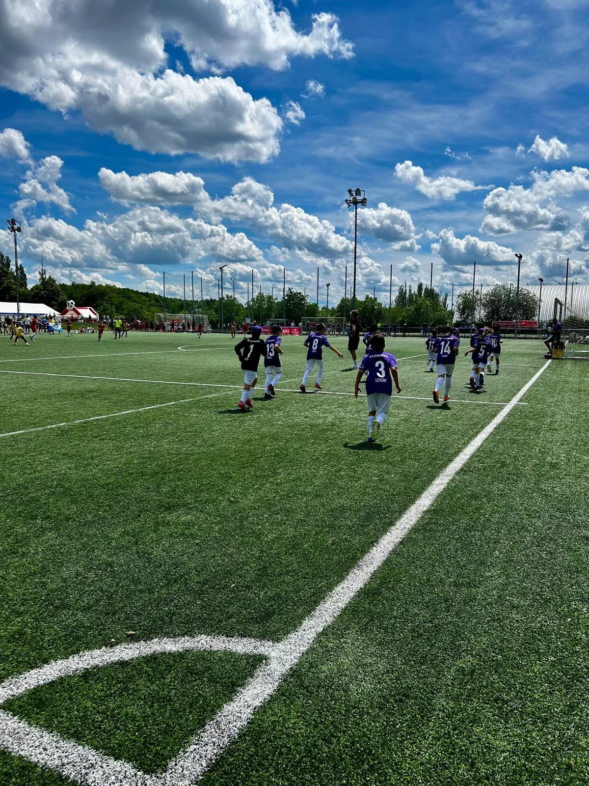 Spielfeld mit Mannschaften beim Aufwärmen, Zuschauer am Spielfeldrand vor dem Vereinsheim